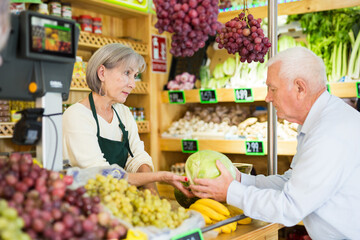 Senior saleswoman standing at counter in greengrocer and serving man customer who purchasing fresh cabbage.