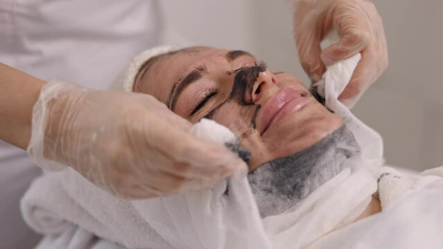Client treatment, Organic skincare, Deep cleansing. Client at beauty salon receives black mask treatment from skincare specialist who wipes off with napkin, providing detox, lifting, kelp rejuvenation