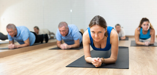 Sportive middle-aged woman doing pilates exercises on gray mat in gym area during workout session. Persons engaging in pilates in fitness room