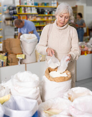Positive interested aged woman making purchases in organic food store, filling plastic bag with white wheat flour from large bag