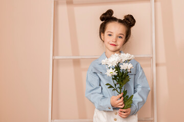 Portrait of a charming little girl, well dressed in a blue shirt and white pants or trousers and with a slight makeup and double bun hair. Isolated on pastel beige background.