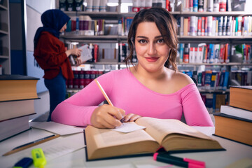 Young and beautiful, student girl studying at the library and taking notes. The girl looks at the camera with a slight smile and a proud look.