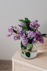 Bouquet of garden lilacs in a glass vase on a wooden bedside table.