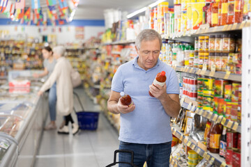 Focused aged man reading labels on bottles with sauces in supermarket, carefully examining ingredients and expiration date while shopping for groceries
