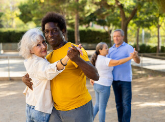 A troupe of cheerful multiracial amateur dancers of different age performing a partner dance outdoors in city park with one couple dancing