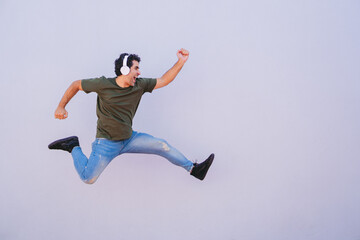 Obraz premium Middle-aged Latin man in profile jumping with clenched fists while wearing headphones on plain background. Copy space.