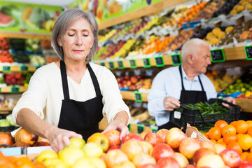 Old lady and man in aprons working in salesroom of greengrocer. Woman setting out goods on shelves, man carrying crate full of green pepper.