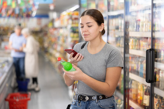 Positive young female customer scanning barcode on bottle of carbonated drink with smartphone while shopping in supermarket, paying for item using mobile app