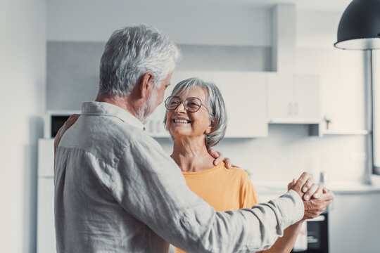 Joyful active old retired romantic couple dancing laughing in living room, happy middle aged wife and elder husband having fun at home, smiling senior family grandparents relaxing bonding together
