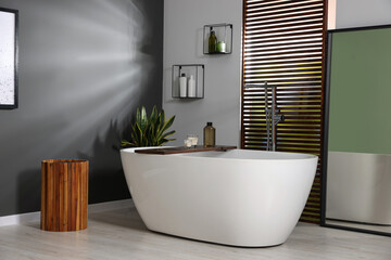 Stylish bathroom interior with ceramic tub, candles and care products on wooden bath tray