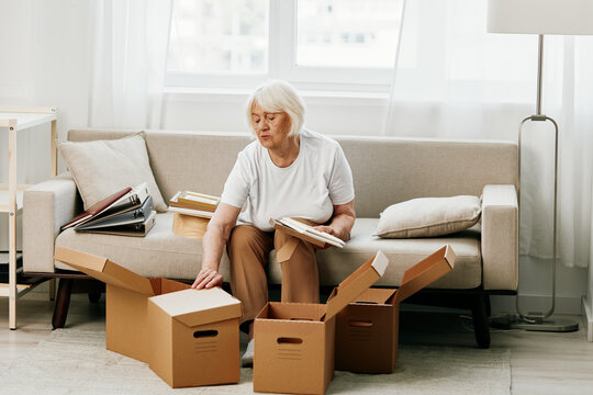 elderly woman sits on a sofa at home with boxes. collecting things with memories albums with photos and photo frames moving to a new place cleaning things and a happy smile. Lifestyle retirement.