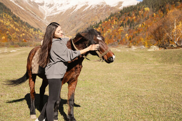 Young woman stroking horse in mountains on sunny day. Beautiful pet