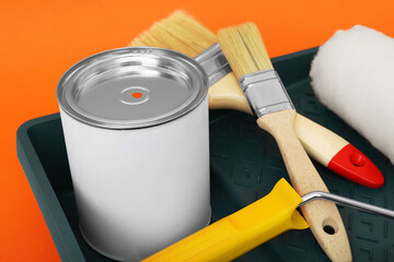 Can of orange paint, brushes, roller and container on color background, closeup