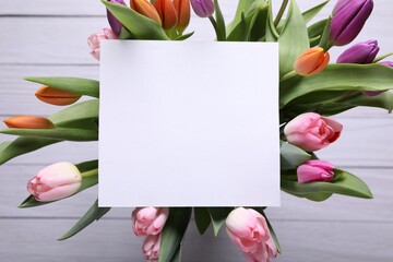 Beautiful bouquet of tulips with blank card on white wooden background, top view. Birthday celebration