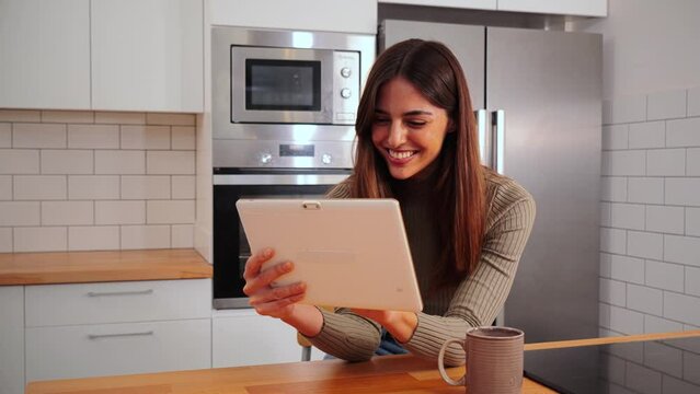 One happy caucasian woman with a tablet device to browse on internet using wireless conection sitting at home kitchen. Isolaten young female smiling and enjoying doing online shopping. Slow motion