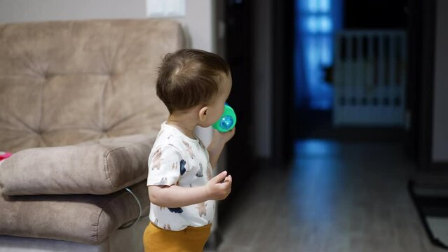 Little child stands in the room drinking water from a bottle. Lovely boy drops the bottle and approaches the camera.