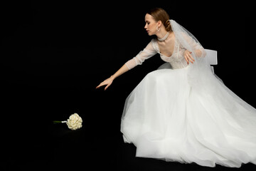 Naklejka premium A beautiful charming girl, the bride in a luxurious and elegant white wedding dress, reaching their hand to a graceful bouquet of white roses lying next to her. Isolated on a black background.