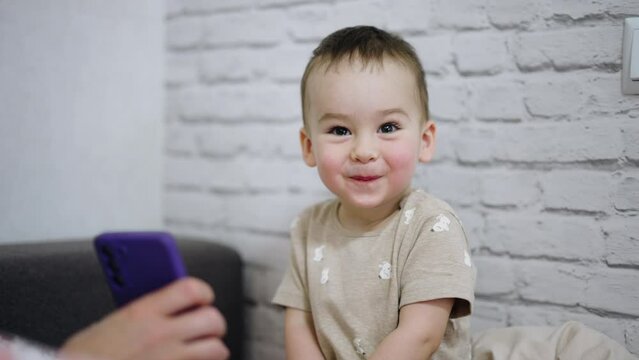 Adorable little boy is amused by something shown on the phone. Cute baby smiles and giggles sweetly. White wall backdrop.