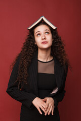 fashion female portrait. Girl, curly girl with black hair in a strict business suit on a red, pink background. High quality photo. Female student rejoices, happy and thoughtful with a book on her head