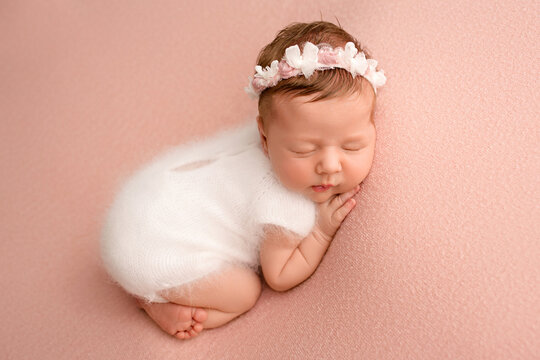 Top view of a newborn baby girl sleeping in a white jumpsuit with a white bandage and a pink flower on her head on a pink background. Beautiful portrait of a little girl 7 days, one week. 