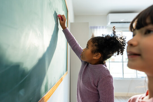 Diverse student writing blackboard in classroom at elementary school.