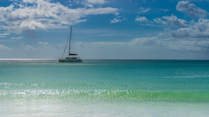 Fototapeta premium Tropical idyll. A white yacht is sailing on a calm turquoise ocean. A mast on a background of blue sky and clouds. The wave rolls towards the shore. Glare on the water. Seychelles.