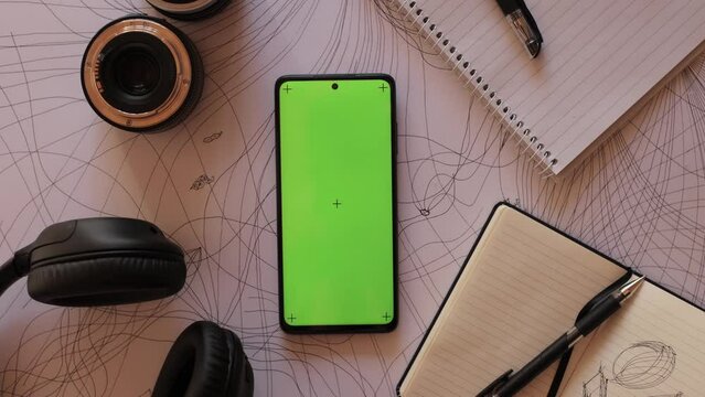 Zoom in Mobile phone with a green screen and motion capture dots, top view, lies on an abstract painting next to a photo lens, a notebook and headphones. Smartphone Application concept for artists