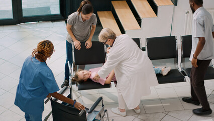 Worried man bringing unconscious girl in hospital lobby, kid fainting and parents asking physician for help. Senior medic and nurse consulting young sick girl in health center. Handheld shot.