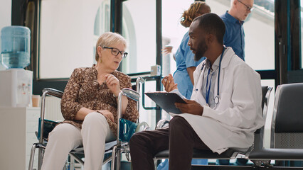 Obraz premium Senior woman with impairment talking to specialist in hospital reception waiting room, consulting wheelchair user in facility lobby. Male doctor taking notes on medical form report.