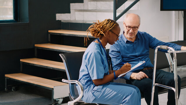 Medical assistant consulting elderly man with walking frame, sitting in hospital reception lobby. Nurse talking to old person in waiting room at health center, filling in clinical checkup report.
