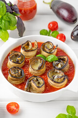 Eggplant rolls with cheese in tomato sauce in a white ceramic baking form on a white wooden background. Vegetarian healthy dish. Selective focus, top view.