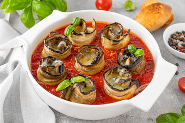 Eggplant rolls with cheese in tomato sauce in a white ceramic baking form on a gray concrete background. Vegetarian healthy dish. Selective focus, top view.