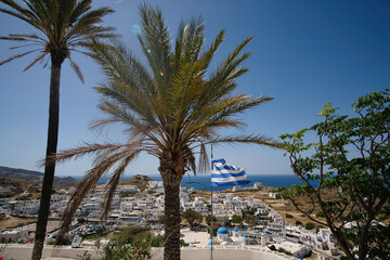 The greek national flag waving in the wind at the top of the village of Ios in Greece surrounded by palm trees