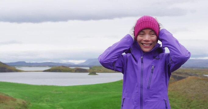 Woman adjusting knit hat wearing hardshell jacket and gloves smiling happy enjoying rugged Iceland nature landscape on hike in classic icelandic weather