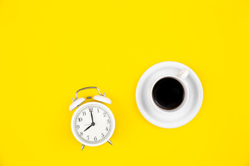 Alarm clock and a cup of coffee on a yellow background, top view.