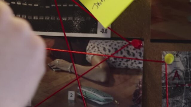 A detective's hand stretches a string between pins on an investigative board with crime scene photos and notes. Close up tracking shot