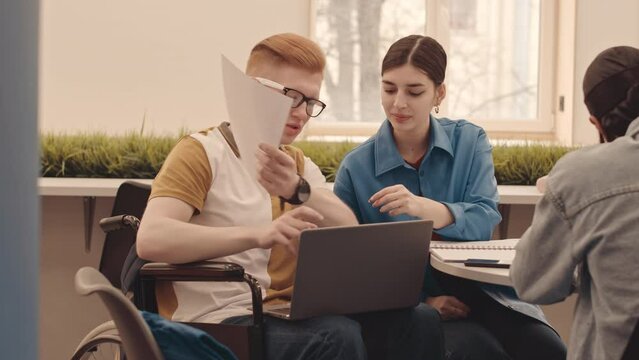 Medium slowmo of Caucasian male university student in wheelchair discussing group project with fellows in coworking space