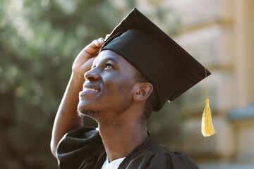 Portrait closeup of afro american student in graduation mantle and hat standing outdoors and with...