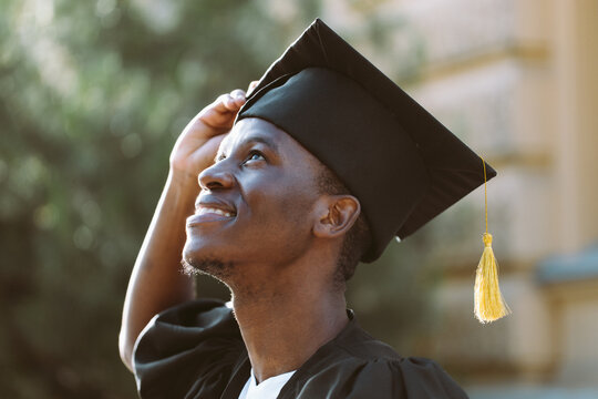 Portrait closeup of afro american student in graduation mantle and hat standing outdoors and with pleasure looking up. Job search, graduate from university, start in life, thoughts about future.