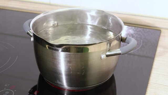 Stationary video of transparent boiling water in stainless steel pot without lid, with steam vapor on black inductive stove panel. Soup preparation, easy cooking at home. Modern kitchenware tools