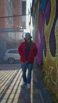 Vertical Video, Stylish Guy in a Cap Stands on background of a Wall Painted with Graffiti. Modern Male Rapper on a City Street in an Urban Style in the Light Sun. Slow motion.