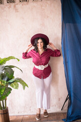A young girl stylist with dark hair smiles in a trendy burgundy color shirt and light trousers and a wide-brimmed burgundy hat in a bright studio with green living plants in pots