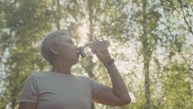 Low angle slowmo of senior Asian woman drinking water from sports bottle while jogging outdoors in park on sunny day