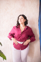 A young girl stylist with dark hair smiles in a trendy burgundy color shirt and light trousers in a bright studio with green living plants in pots