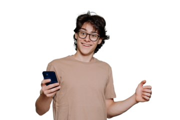 Excited teen in glasses toothy smiles holds phone, amazed by message. Young brunet guy in beige t-shirt, glasses received great news, entered university. American boy isolated, transparent background