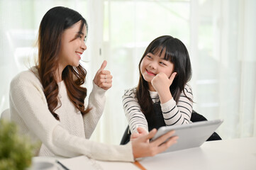 Cheerful Asian mom and her daughter spending time together at home, using tablet