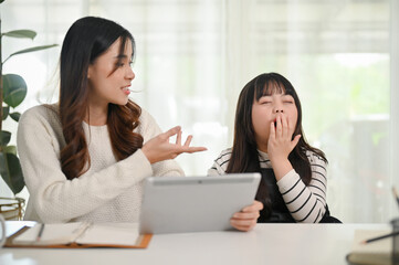 Adorable little Asian girl yawning while her mom is teaching math on tablet at home.