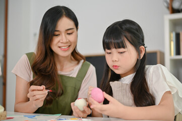 Kind Asian mom helping her little daughter paint an Easter egg, spending fun time together