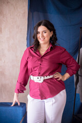 A young stylist girl with dark hair smiles in a trendy burgundy white jacket and light trousers in a light studio with green live plants in pots and a rack with clothes