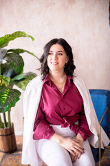 Fototapeta na wymiar A young girl stylist with dark hair smiles in a trendy white jacket, a burgundy color shirt and light trousers in a light studio with green living plants in pots 